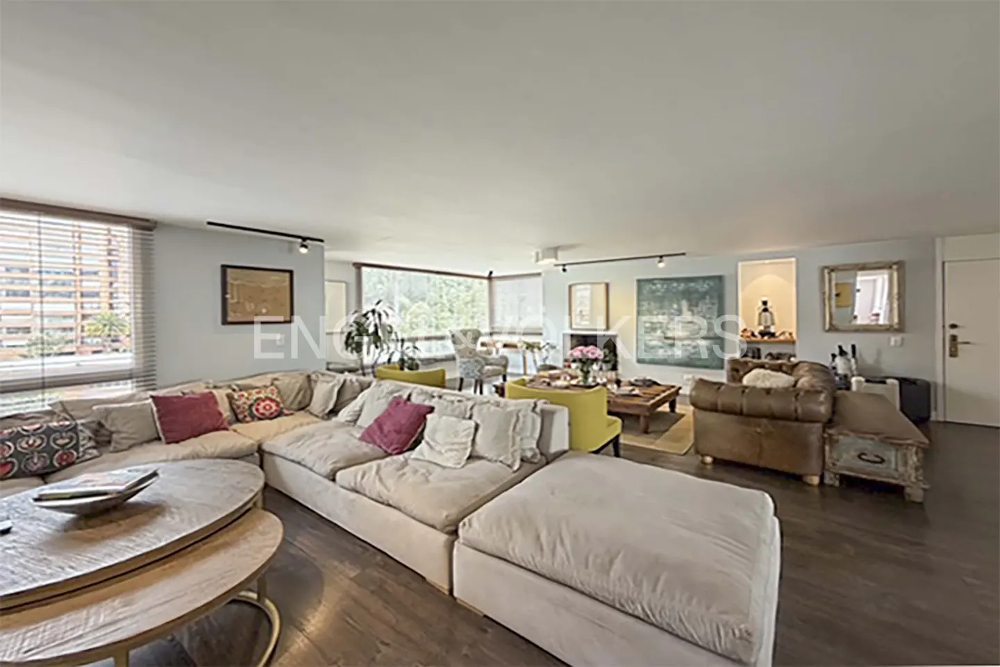Wide shot of a living room with a large beige sectional sofa, wooden floors, and large windows with blinds.