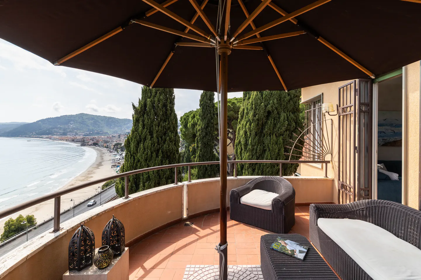 Balcony with brown umbrella, wicker furniture, and ocean view. Tall green trees and a sandy beach are visible in the background.
