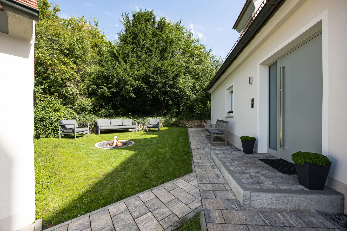 Exterior view of a white house with a gray stone walkway leading to the front door. A green lawn features a fire pit and outdoor seating.