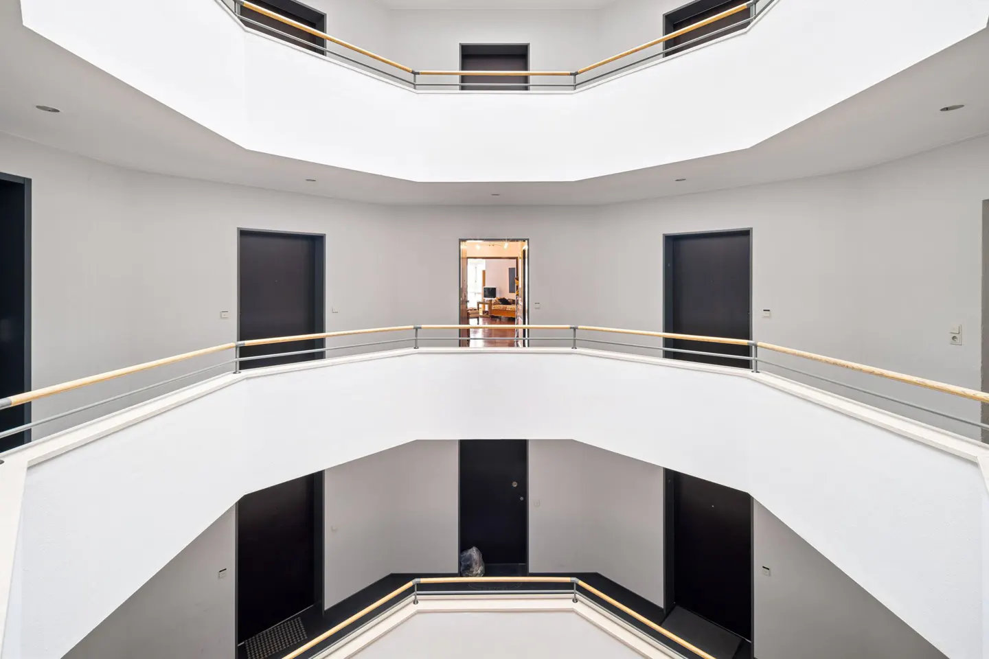 Interior view of a three-story building with white walls, black doors, and wooden handrails.