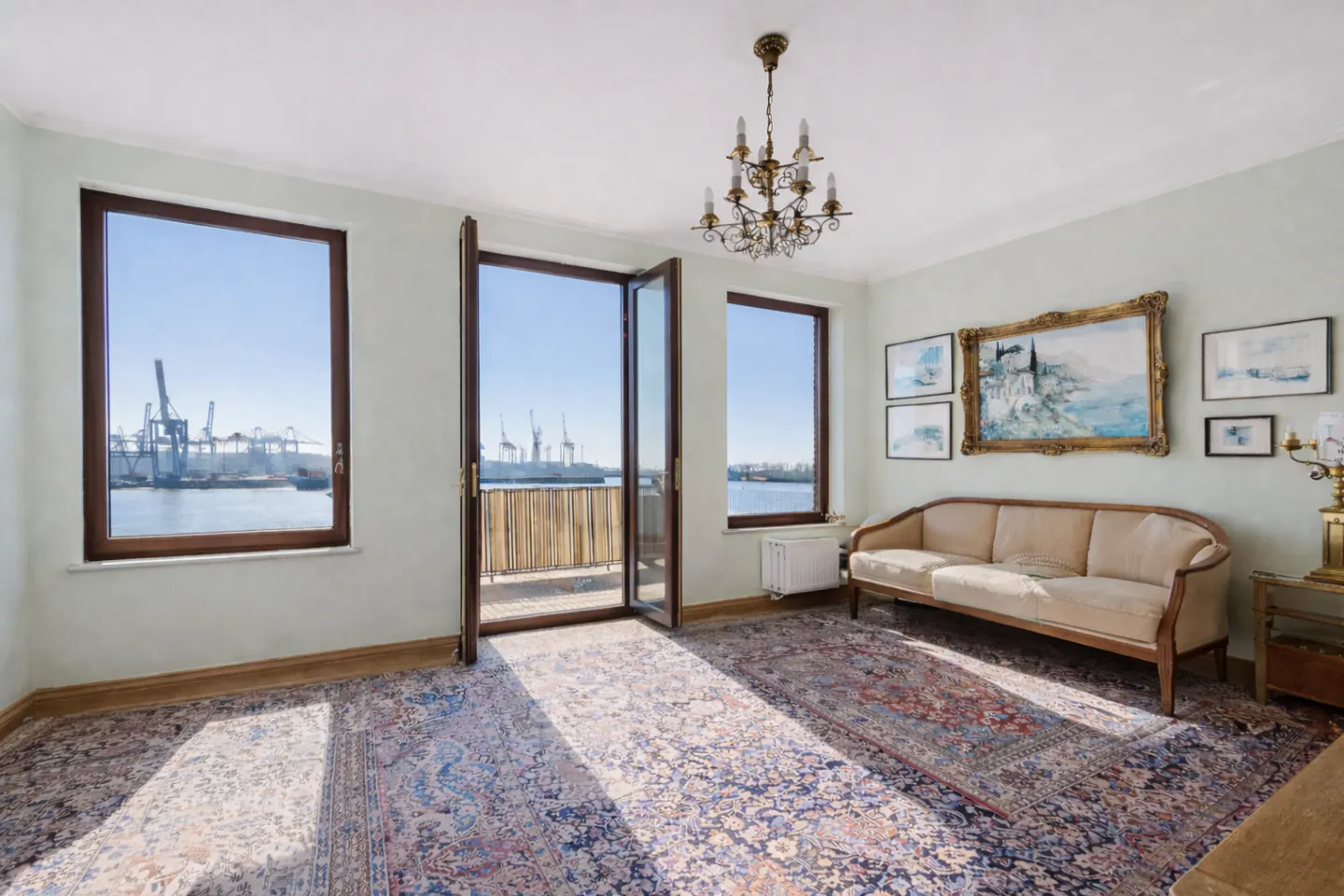 Bright living room with harbor view. Beige sofa, ornate rug, and gold chandelier. Windows show cranes and water.
