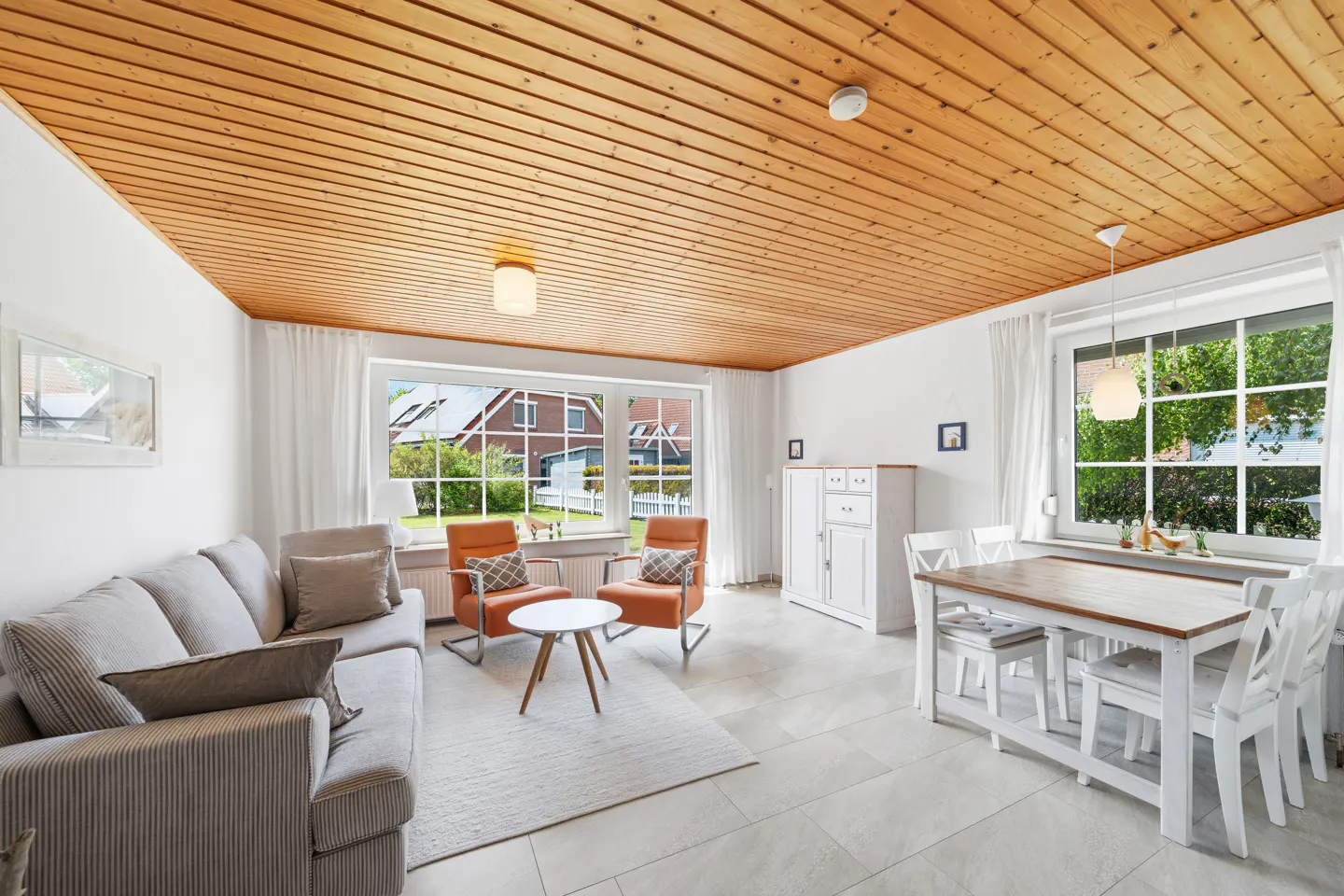 Bright living room with wood ceiling, gray sofa, orange chairs, white table and chairs, and large windows.