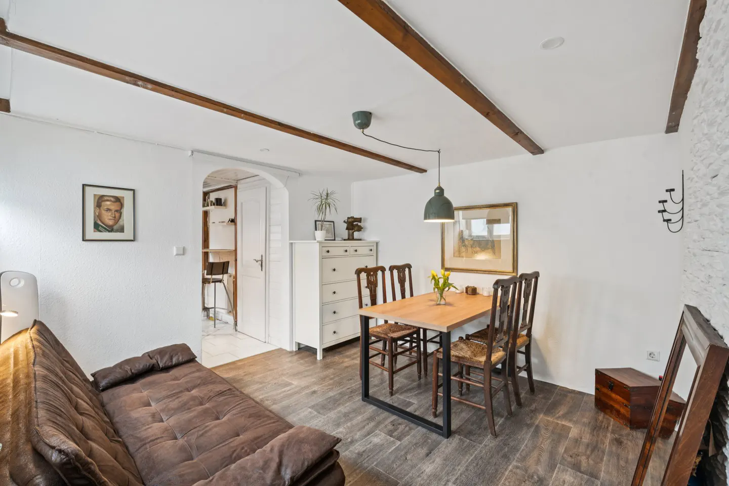 Living room with brown sofa, wooden dining table with chairs, white dresser, and exposed wooden beams on a white ceiling.