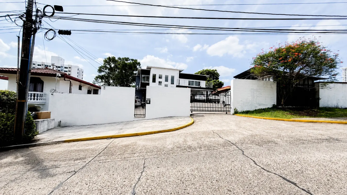 Exterior view of a gated property with white walls and a modern house in the background.