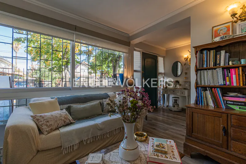 Living room with a beige sofa, large window, and a wooden bookcase filled with books. A vase of flowers sits on a coffee table.