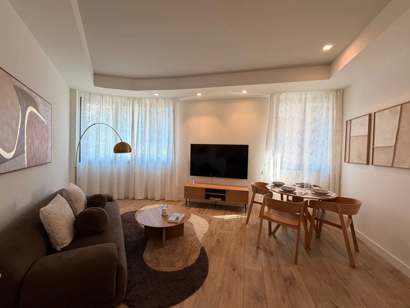 Living room with brown sofa, wood floors, dining table, TV, and abstract art. White walls and curtains create a bright, modern space.