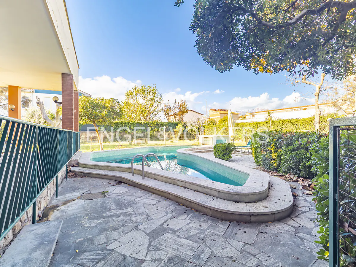 Outdoor pool with turquoise water, surrounded by stone patio and green foliage, under a blue sky. Engel & Volkers logo visible.