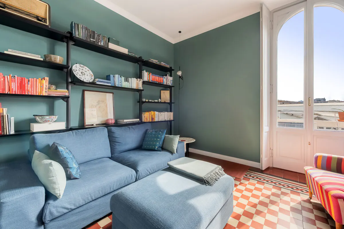 Living room with blue sofa, ottoman, and black shelves filled with books against a teal wall. A white arched door leads to a balcony.