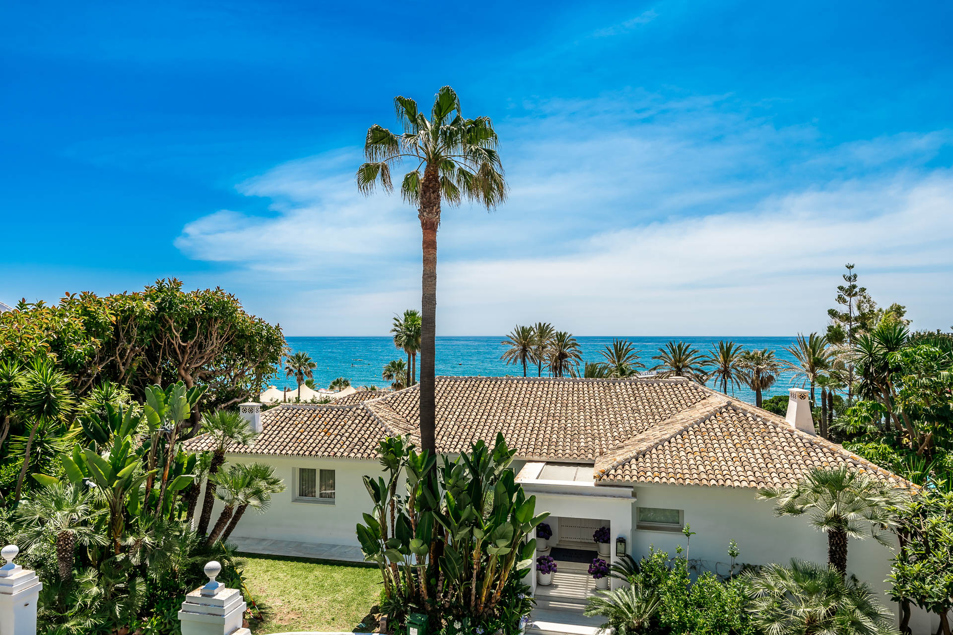 White villa with a tiled roof surrounded by lush tropical gardens and palm trees, overlooking the blue ocean under a sky.