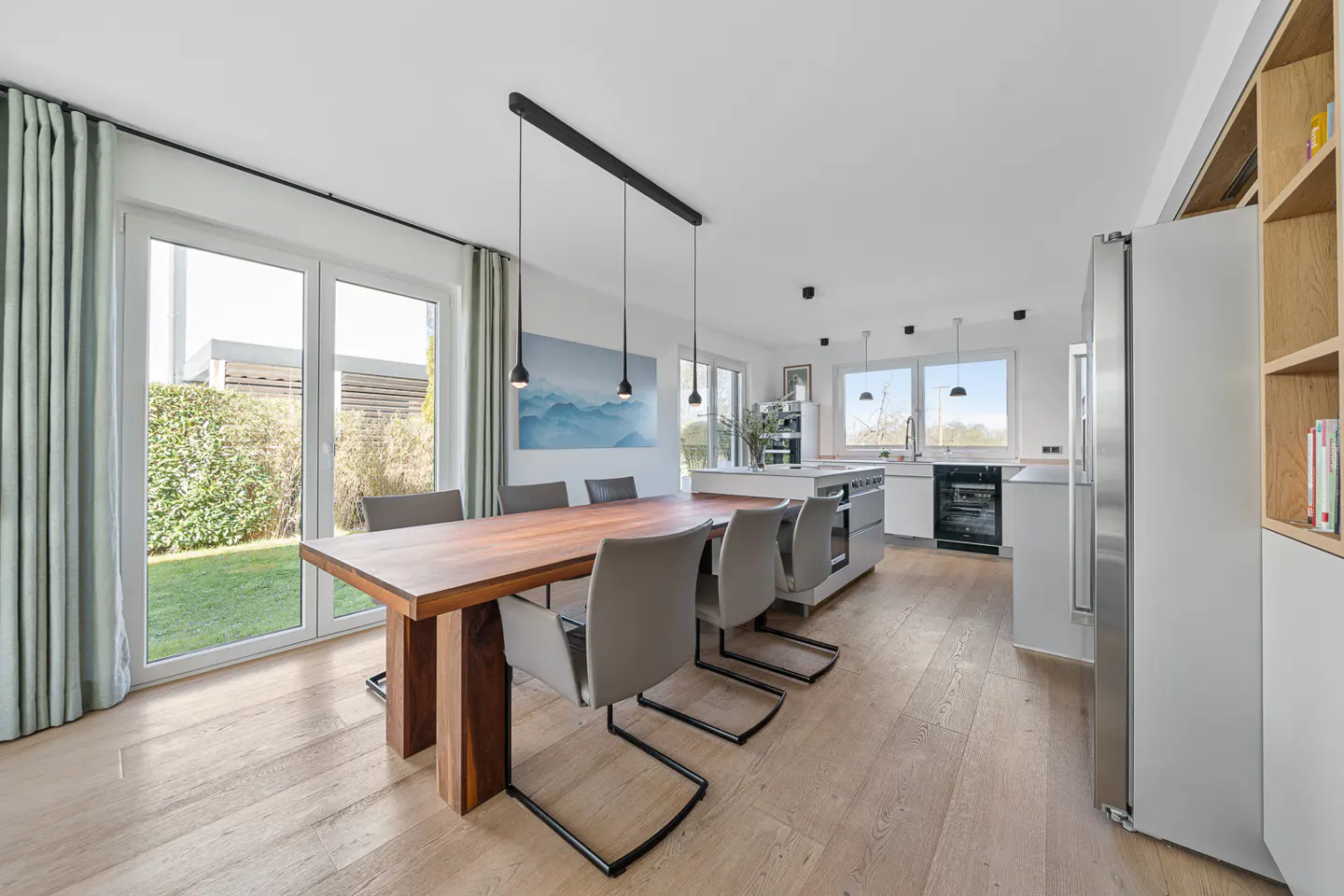 Bright, modern kitchen and dining area with a wood table, gray chairs, and stainless steel appliances. Large windows overlook a green lawn.
