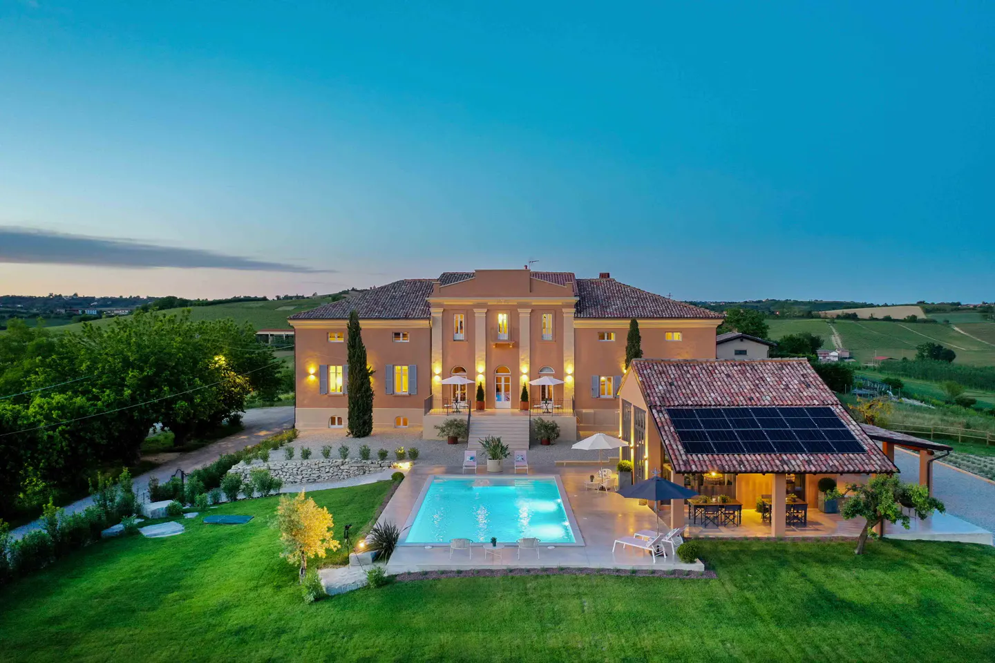 Aerial view of a large, peach-colored villa with a pool, green lawn, and a building with solar panels at dusk.