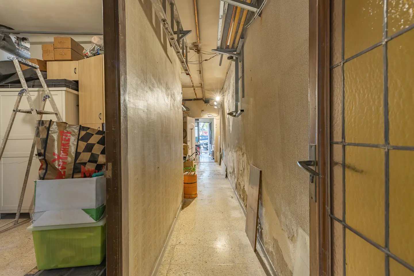 A narrow, cluttered hallway with a tiled floor, leading to a bright doorway, with storage boxes and a ladder on the left.