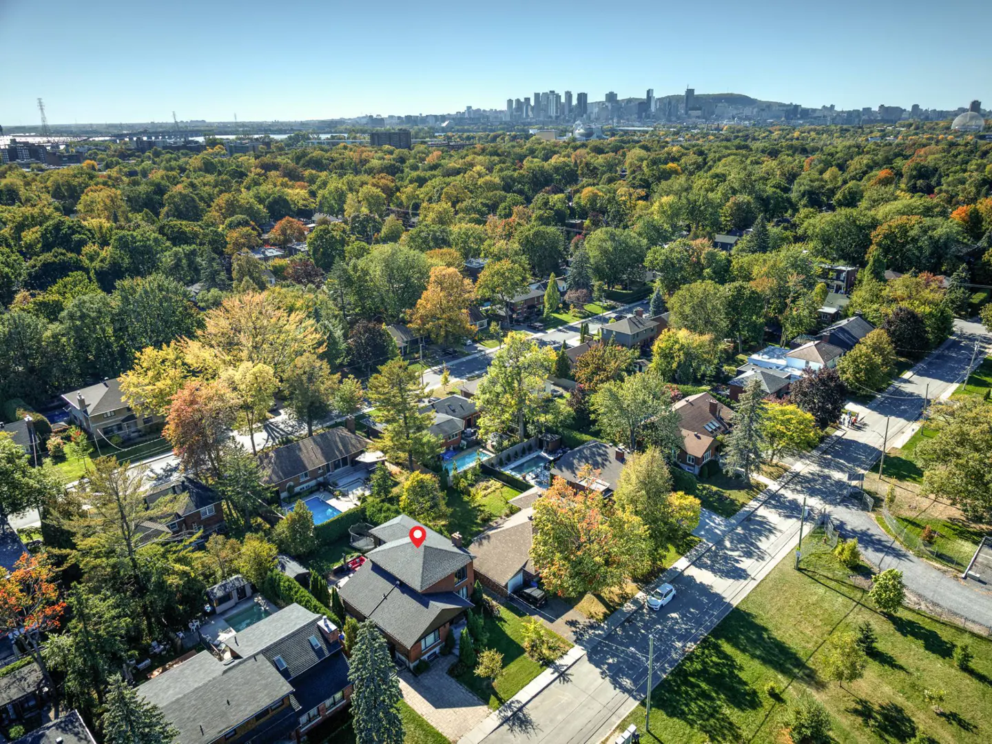 Aerial view of a suburban neighborhood with lush green trees and a city skyline in the distance. A red pin marks a house.