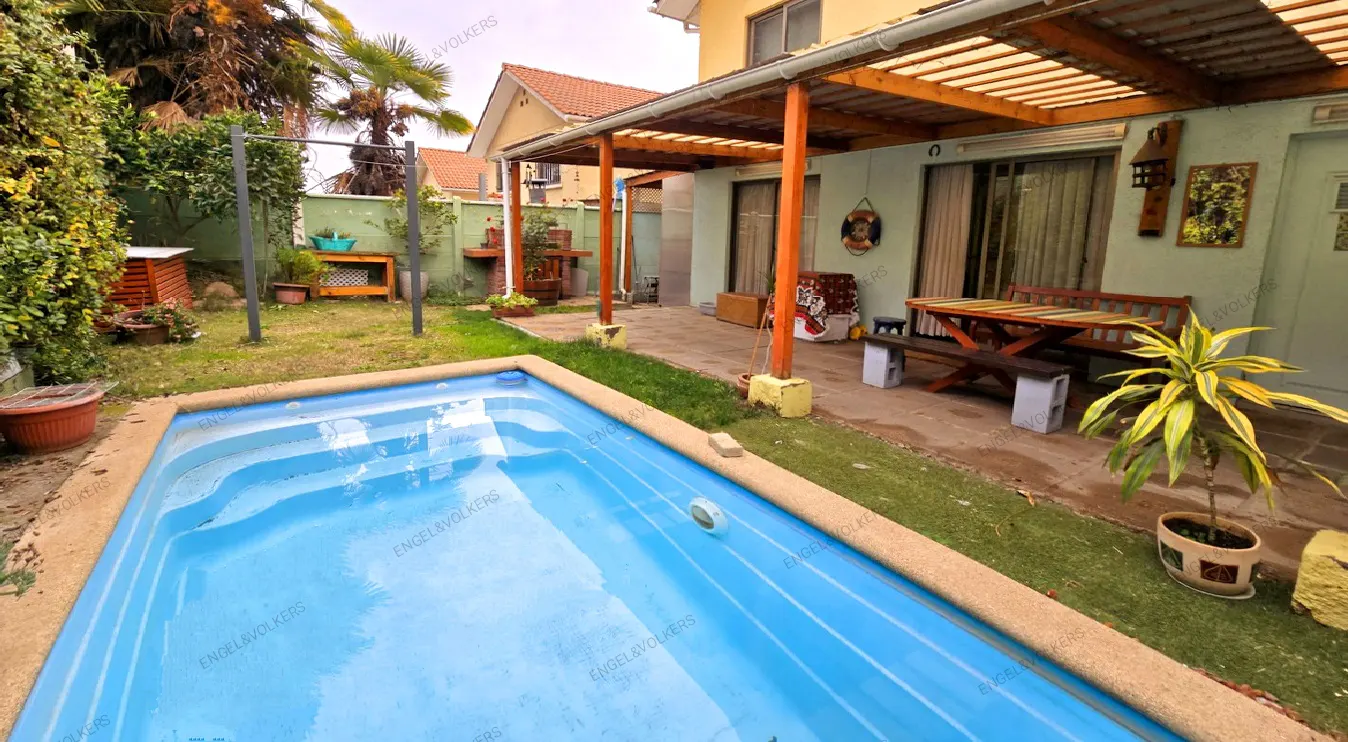 A backyard with a blue pool, green grass, and a covered patio with a wooden table and benches.