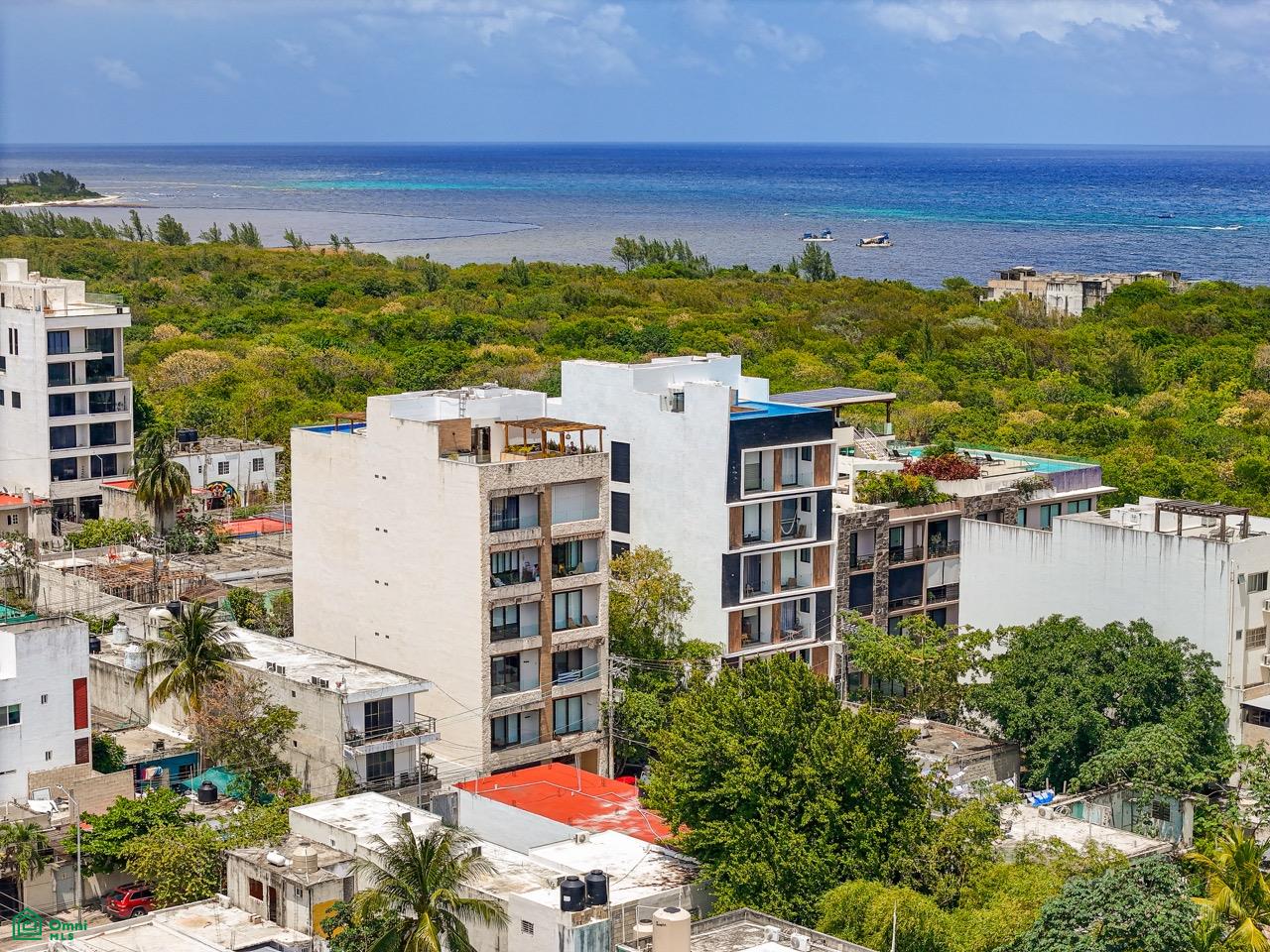 Aerial view of modern white apartment buildings surrounded by lush green trees with the blue ocean in the background. [AI-generated text]