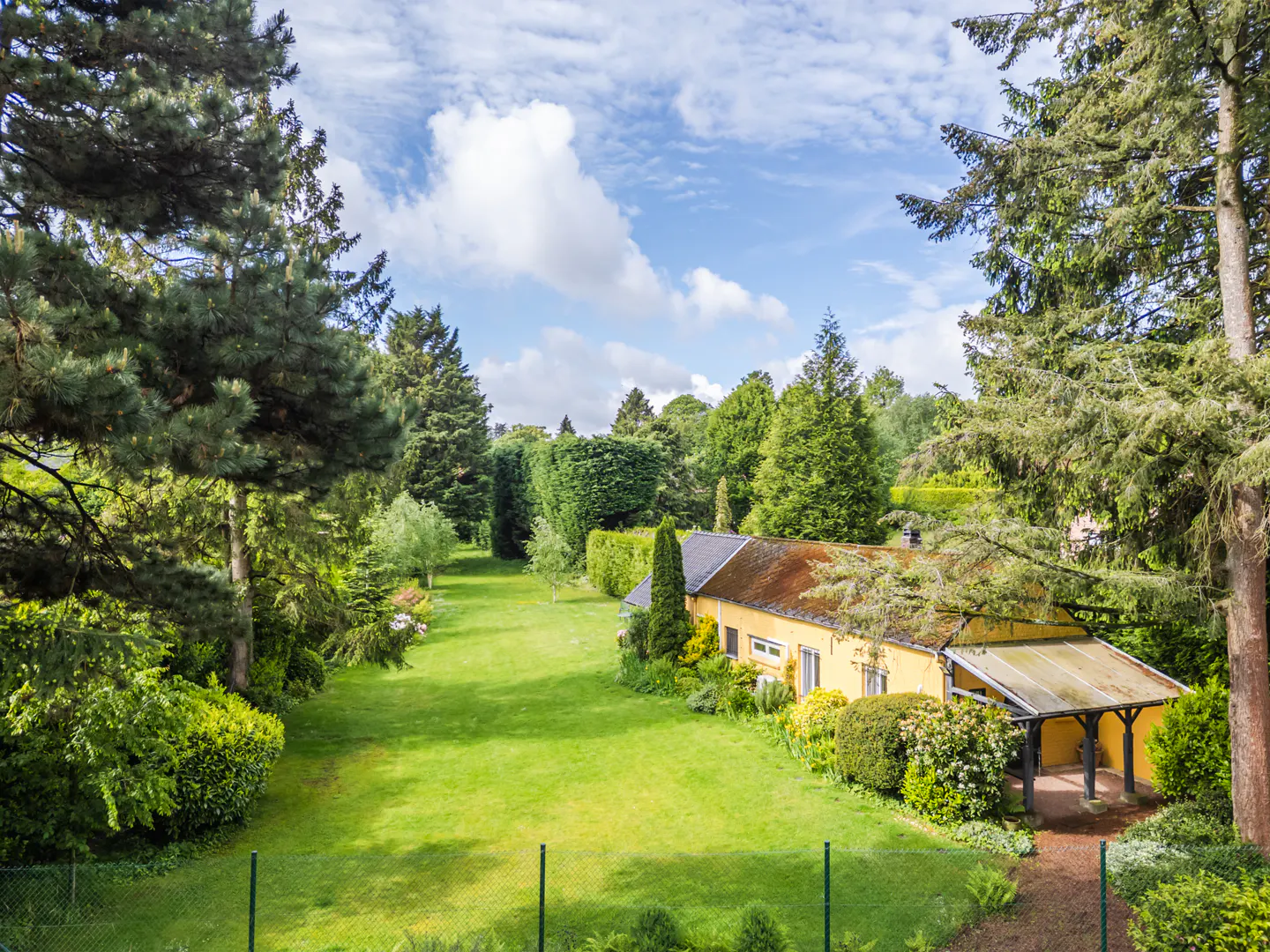 Aerial view of a yellow house with a brown roof, surrounded by green trees and a large lawn under a blue sky.