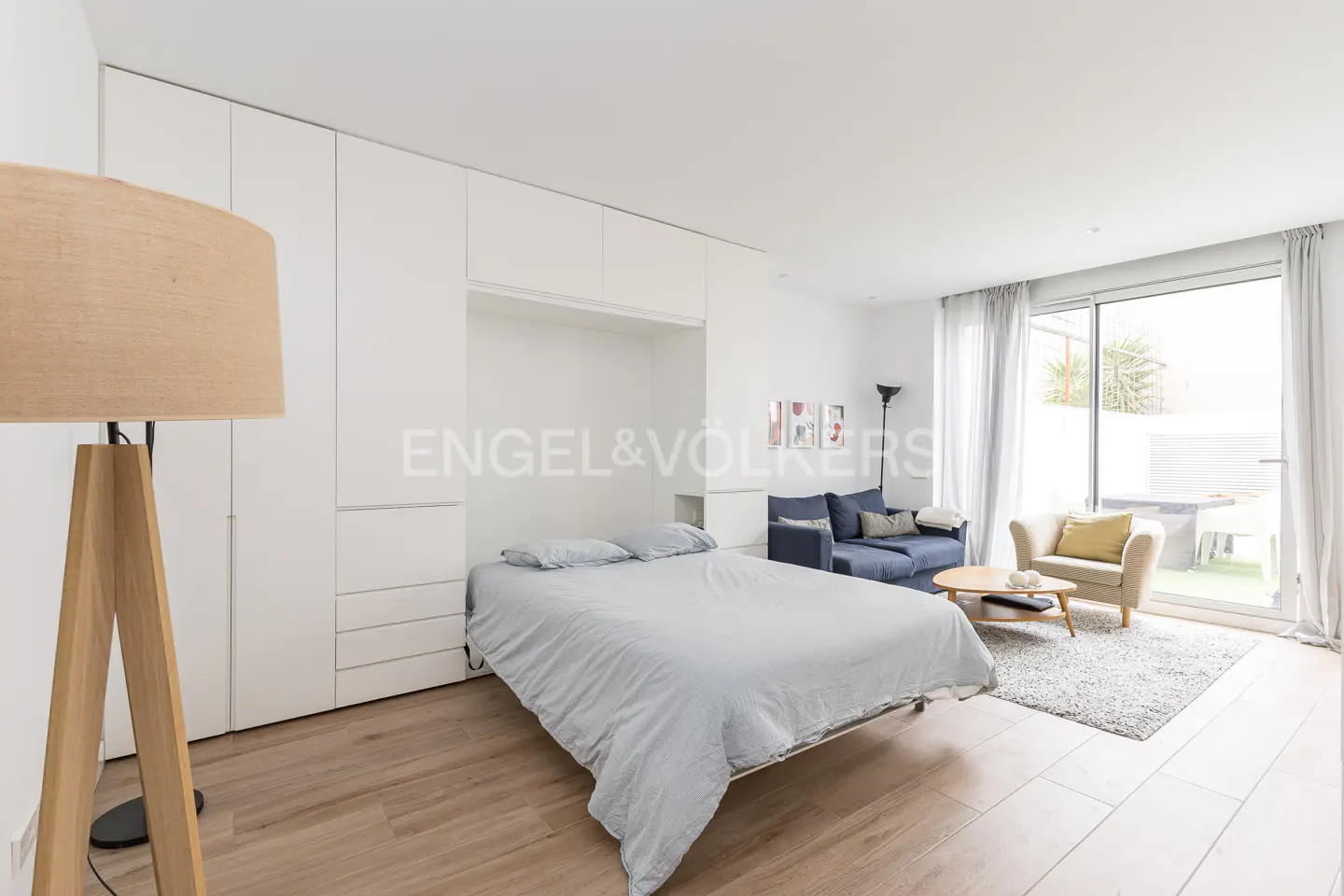 Bright studio apartment with a pull-down bed, white built-in cabinets, a blue sofa, and a sliding glass door to a patio.