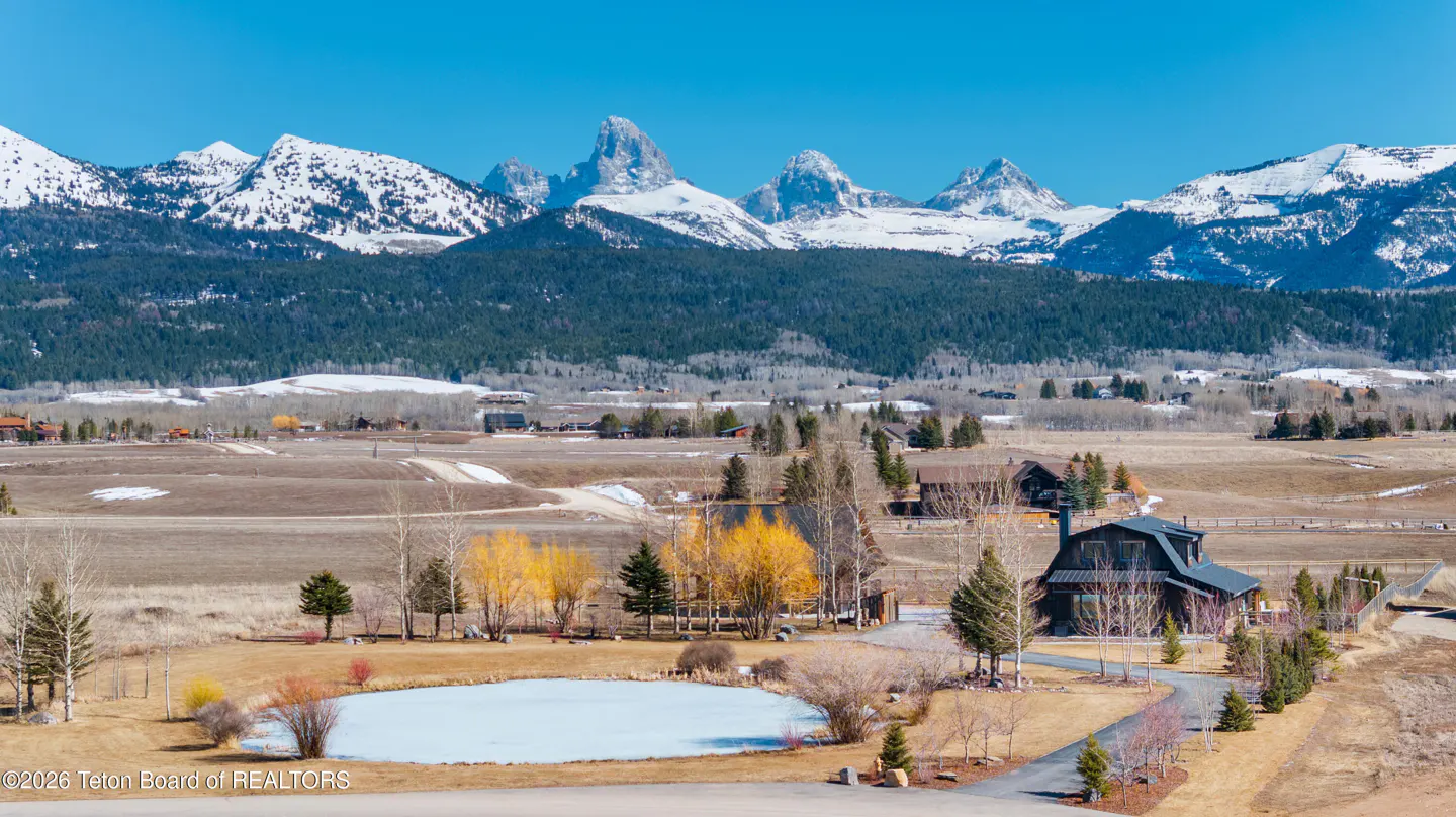 Scenic view of a rural property with snow-capped mountains, a pond, and a modern barn-style house under a clear blue sky.