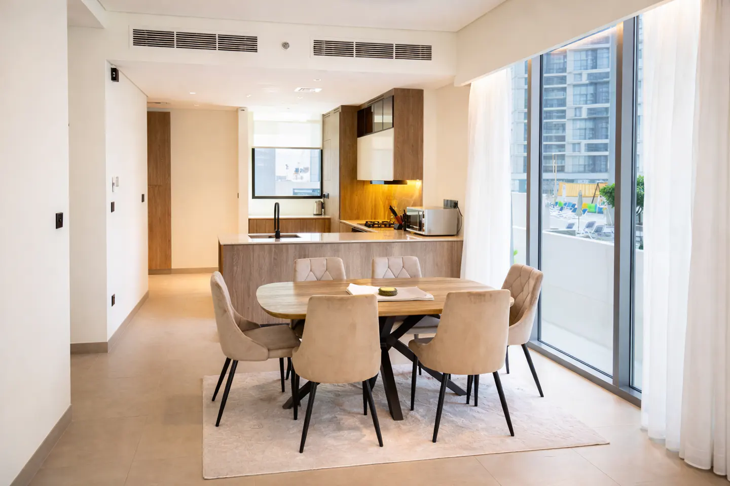 Open-concept apartment with a dining table and kitchen. Six beige chairs surround a wooden table. Large windows offer a city view.