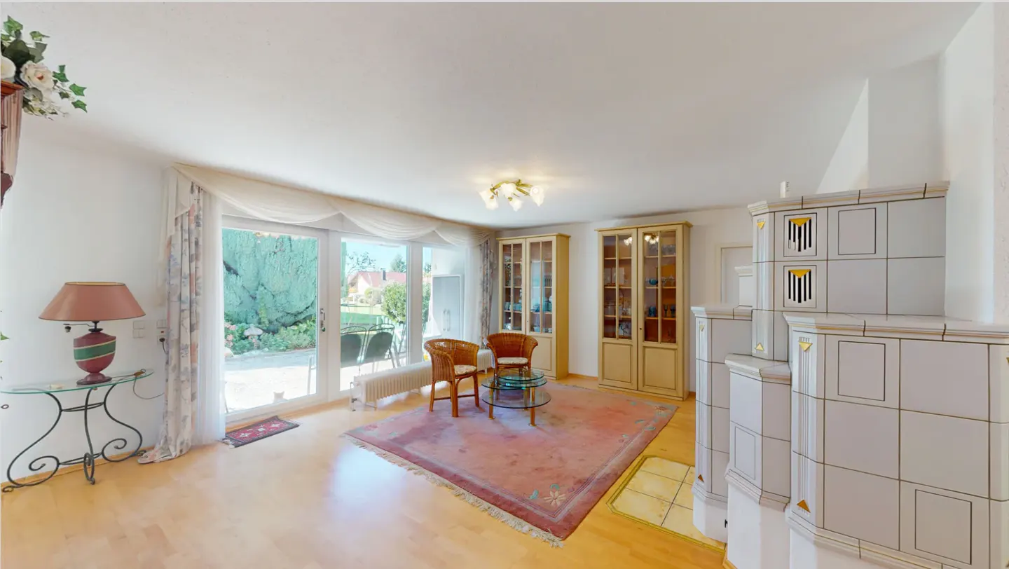 Bright living room with wood floors, a pink rug, and wicker chairs. Sliding glass doors lead to a garden. A white tiled fireplace stands in the corner.