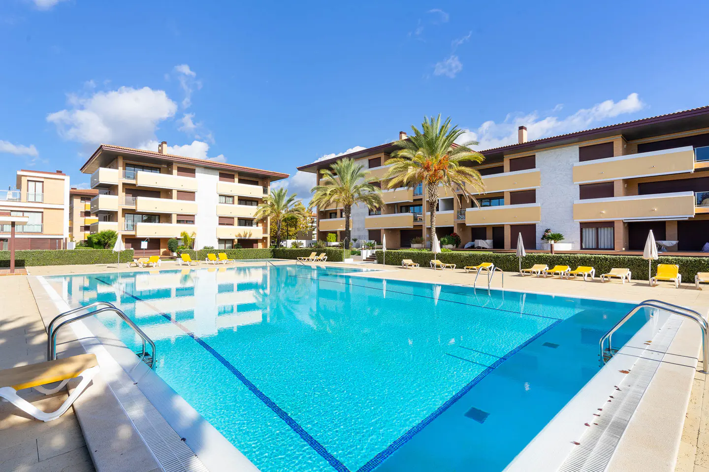 Outdoor pool with clear blue water, surrounded by apartment buildings, palm trees, and yellow lounge chairs under a sunny sky.