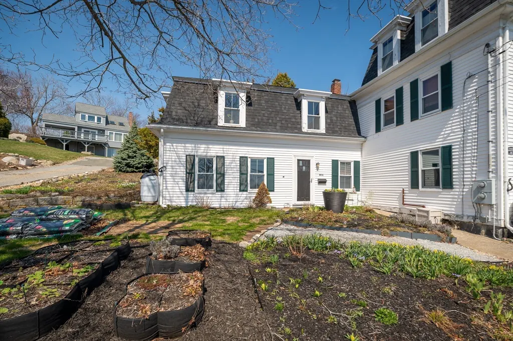Exterior view of a white two-story house with green shutters and a black roof. A garden is in the foreground.