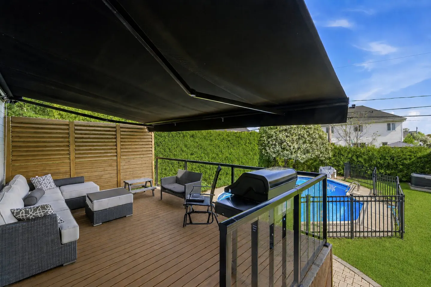 A backyard deck with a black awning, wicker furniture, a grill, and a fenced-in pool on a sunny day.