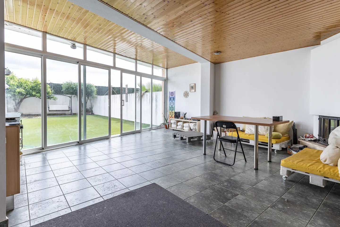 Bright room with wood ceiling, gray tile floor, and large sliding glass doors to a green lawn. A table, chair, and pallet couches furnish the space.