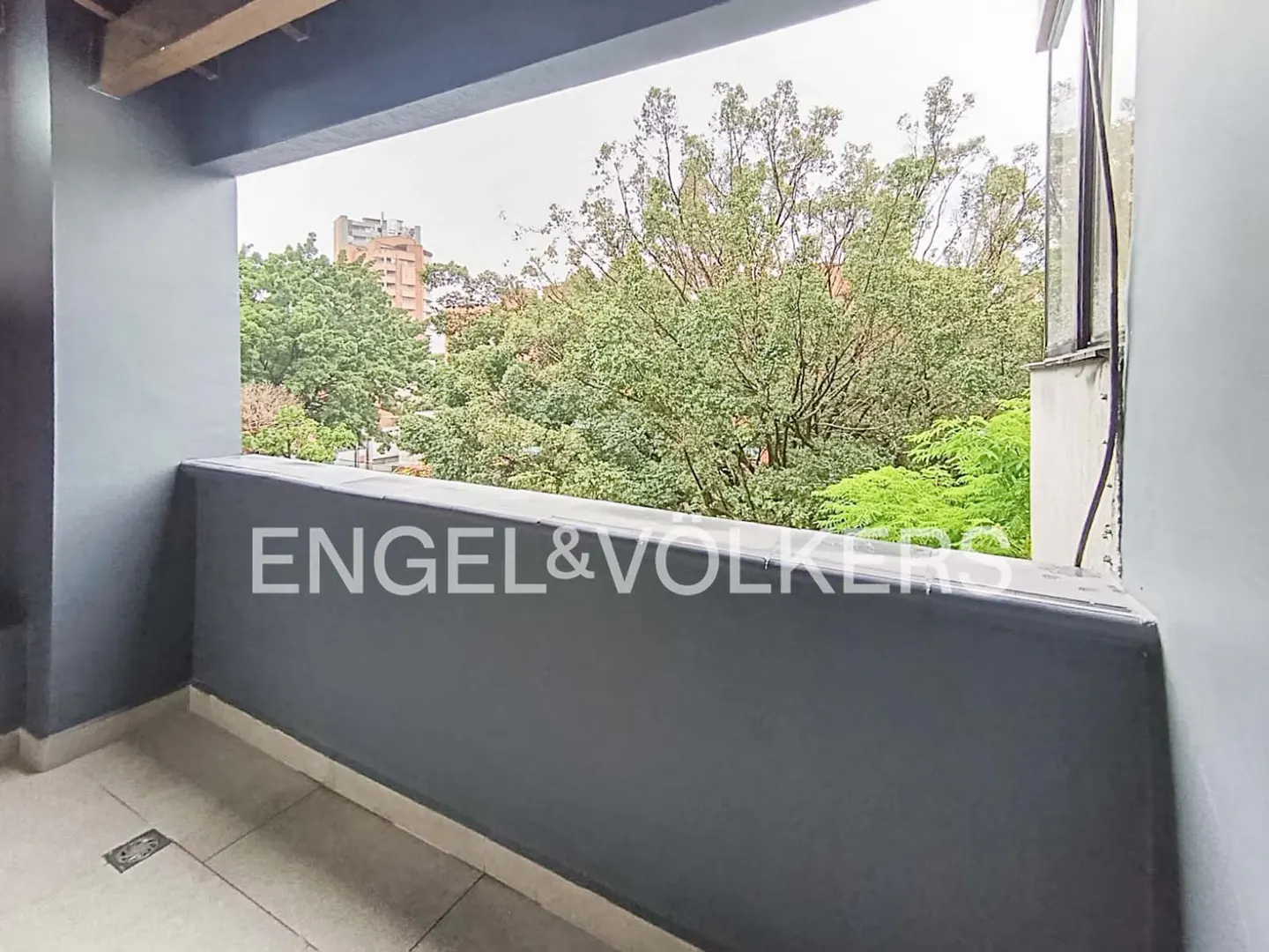 A gray balcony with a view of green trees and a building in the distance. The Engel & Volkers logo is on the balcony wall.
