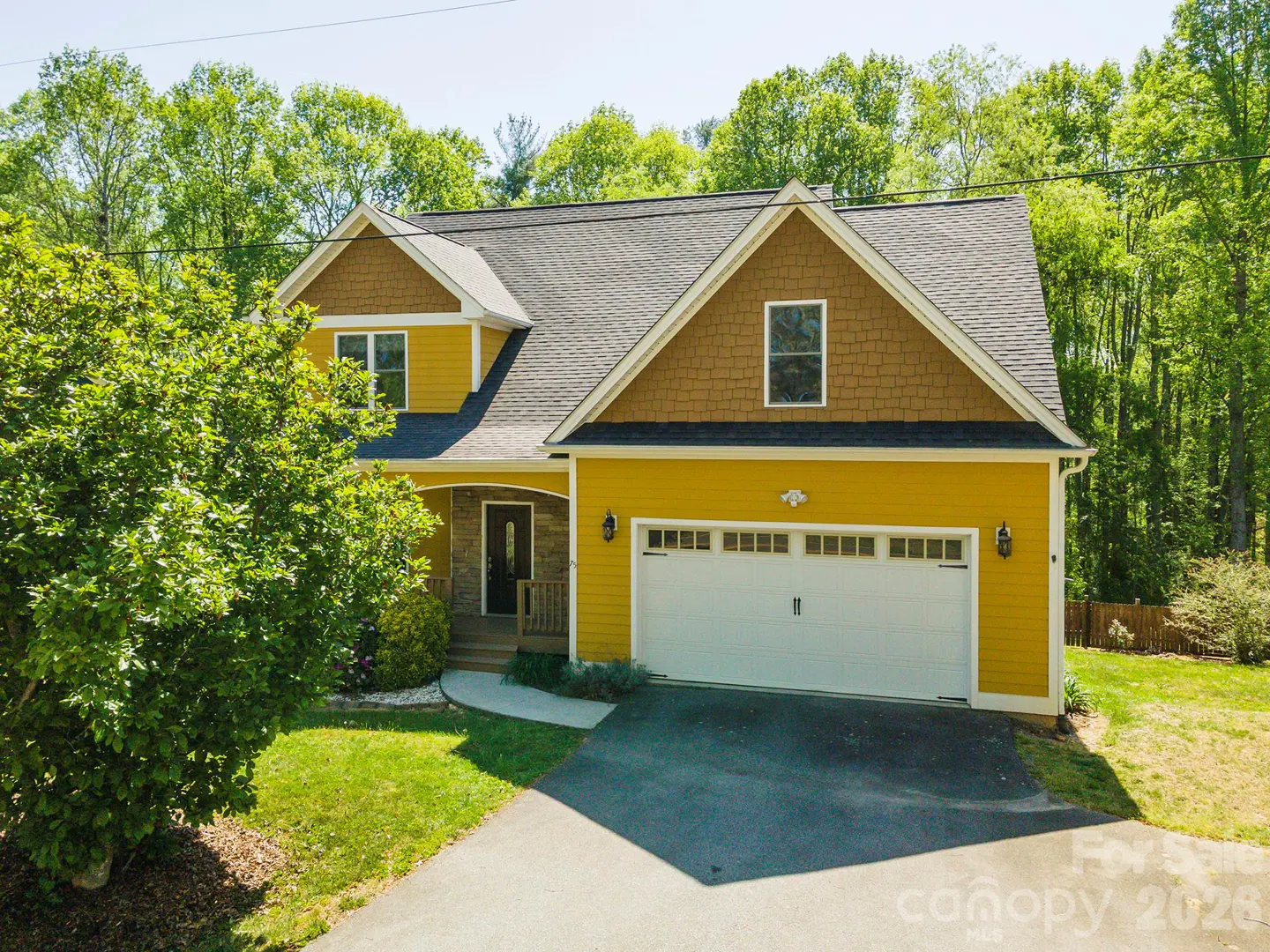 Two-story yellow house with a white garage door, brown trim, and a black roof surrounded by green trees.