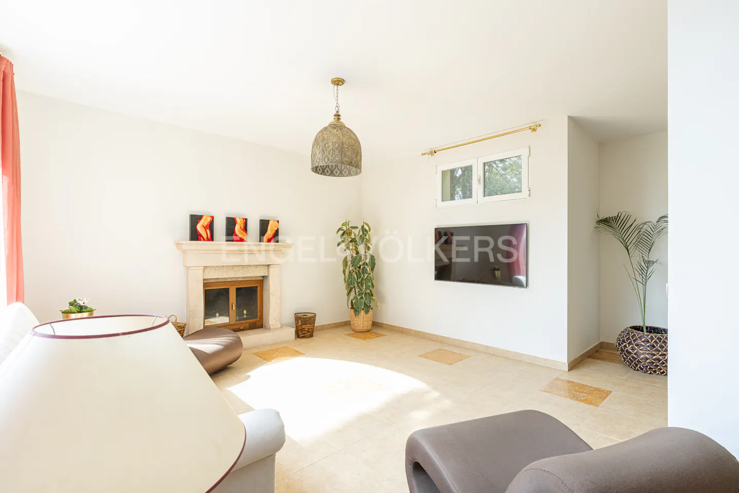 Bright living room with white walls, tile floor, fireplace, TV, and plants. A patterned lamp hangs from the ceiling.