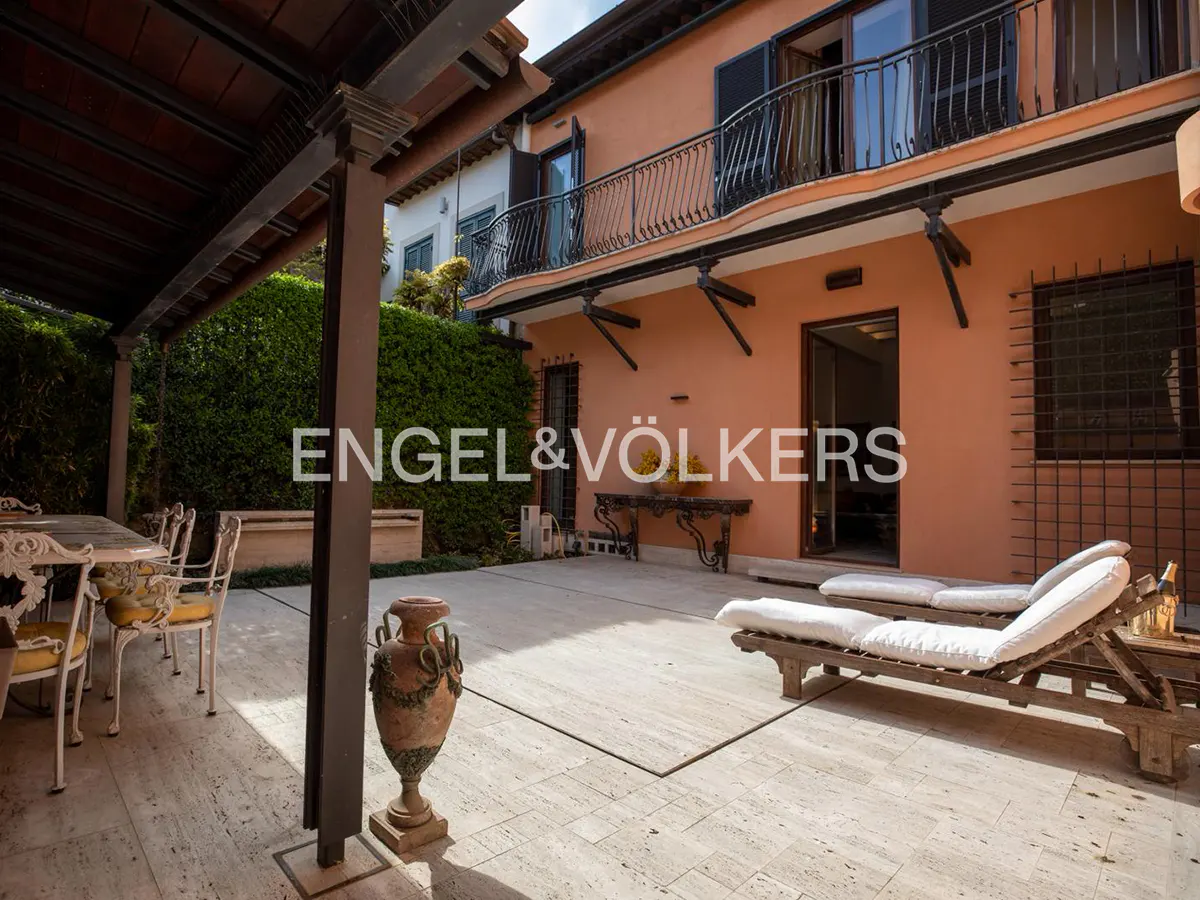 A sunlit stone patio with lounge chairs, a dining set under a pergola, and a terracotta-colored house with a balcony.