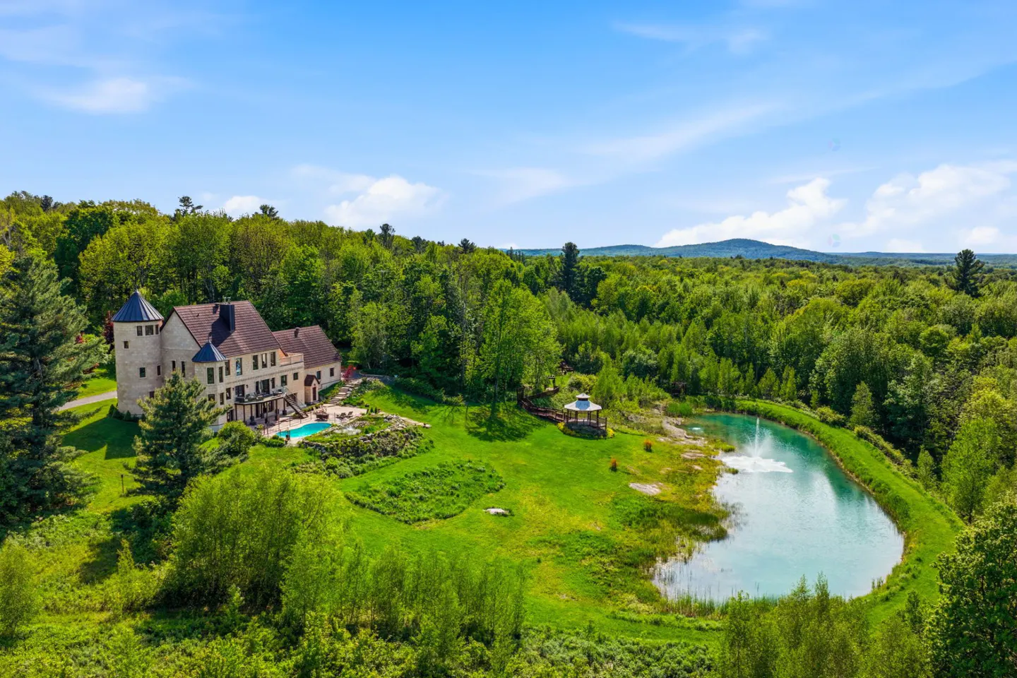 Aerial view of a large stone estate with a pool, gazebo, and pond, surrounded by lush green forest under a blue sky.