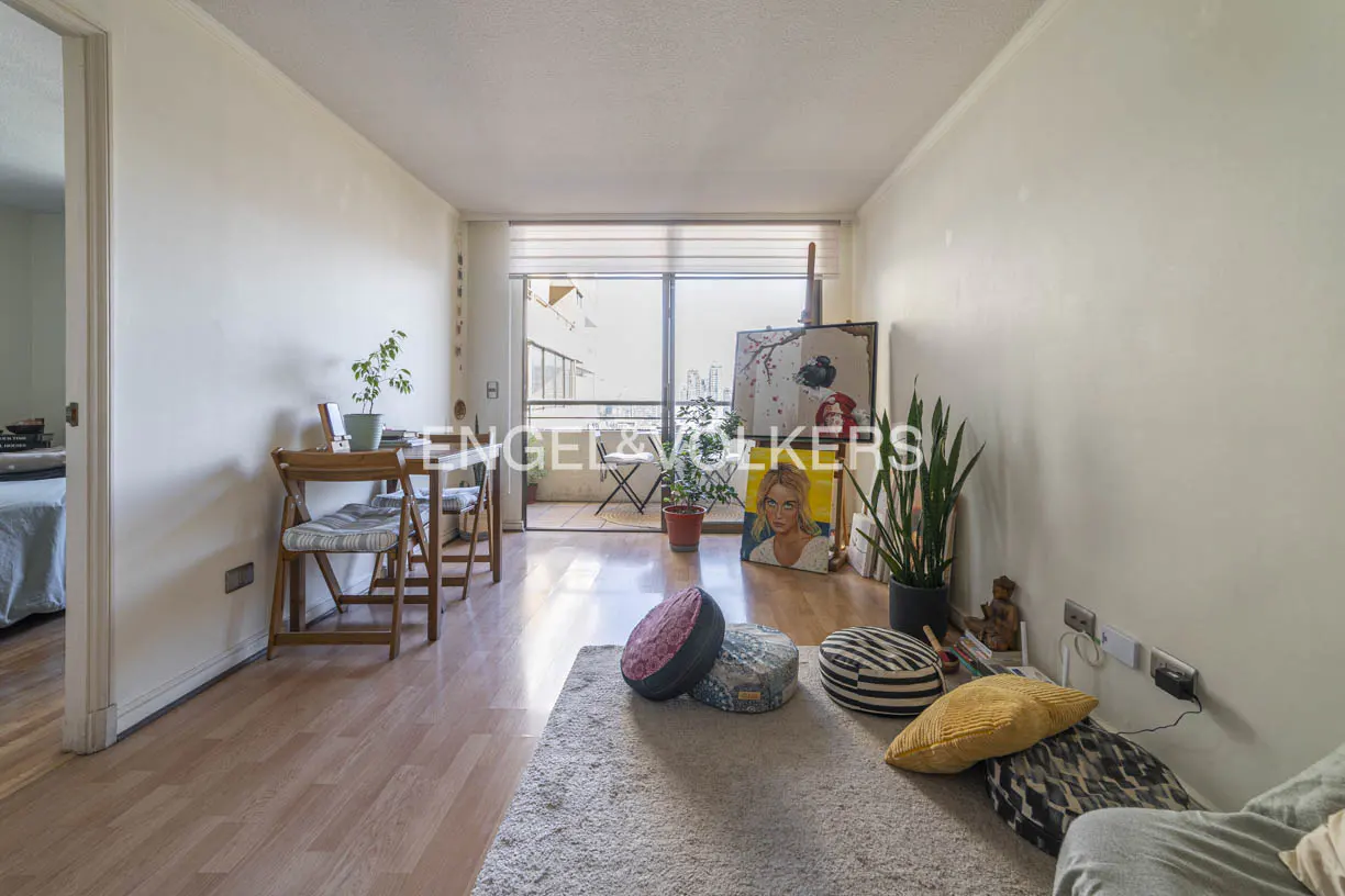 A bright living room with a balcony, wooden floors, and white walls. A table with chairs is on the left, and pillows are on a rug in the foreground.