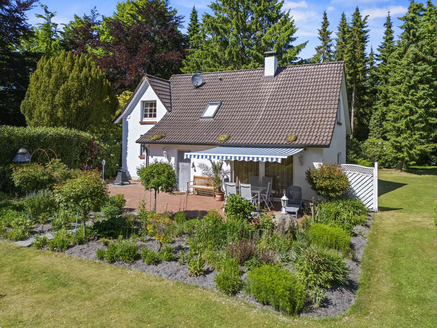 A white house with a brown tiled roof, a blue and white striped awning, and a garden with green plants.