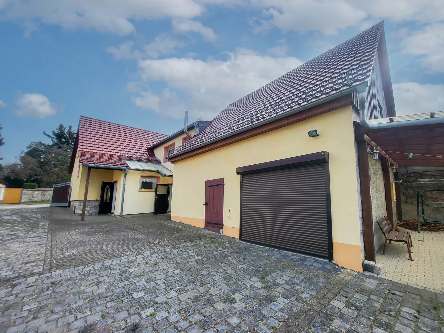 Exterior view of a yellow house with a brown garage door and red roof, set against a blue sky with clouds. Cobblestone driveway.