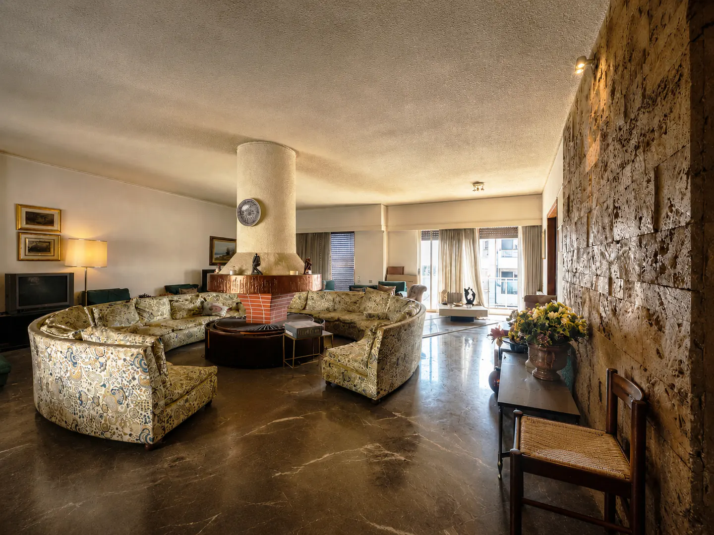 A spacious living room with a round sofa, a cylindrical fireplace, and a textured stone wall. The floor is marble, and natural light streams through the windows.