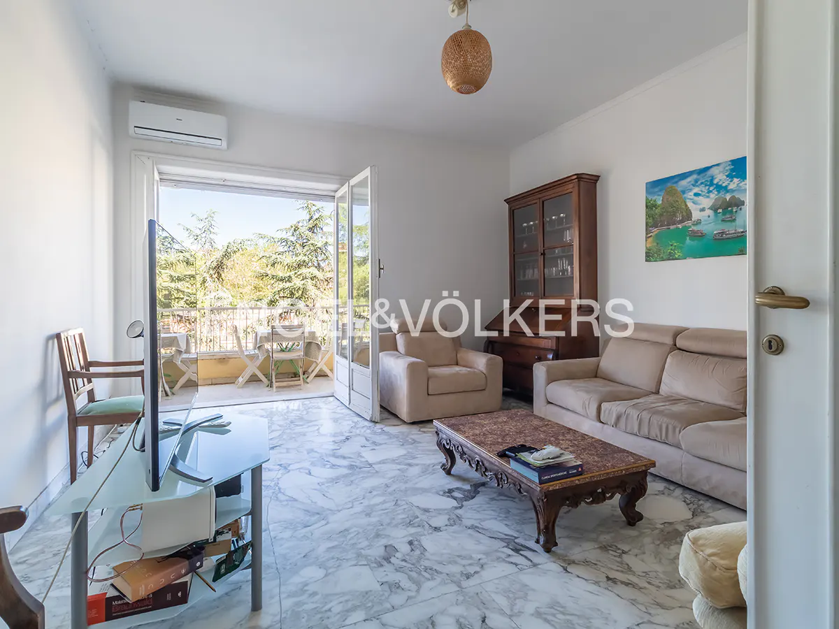 Living room with marble floors, beige sofa and armchair, dark wood cabinet, and balcony with outdoor seating.
