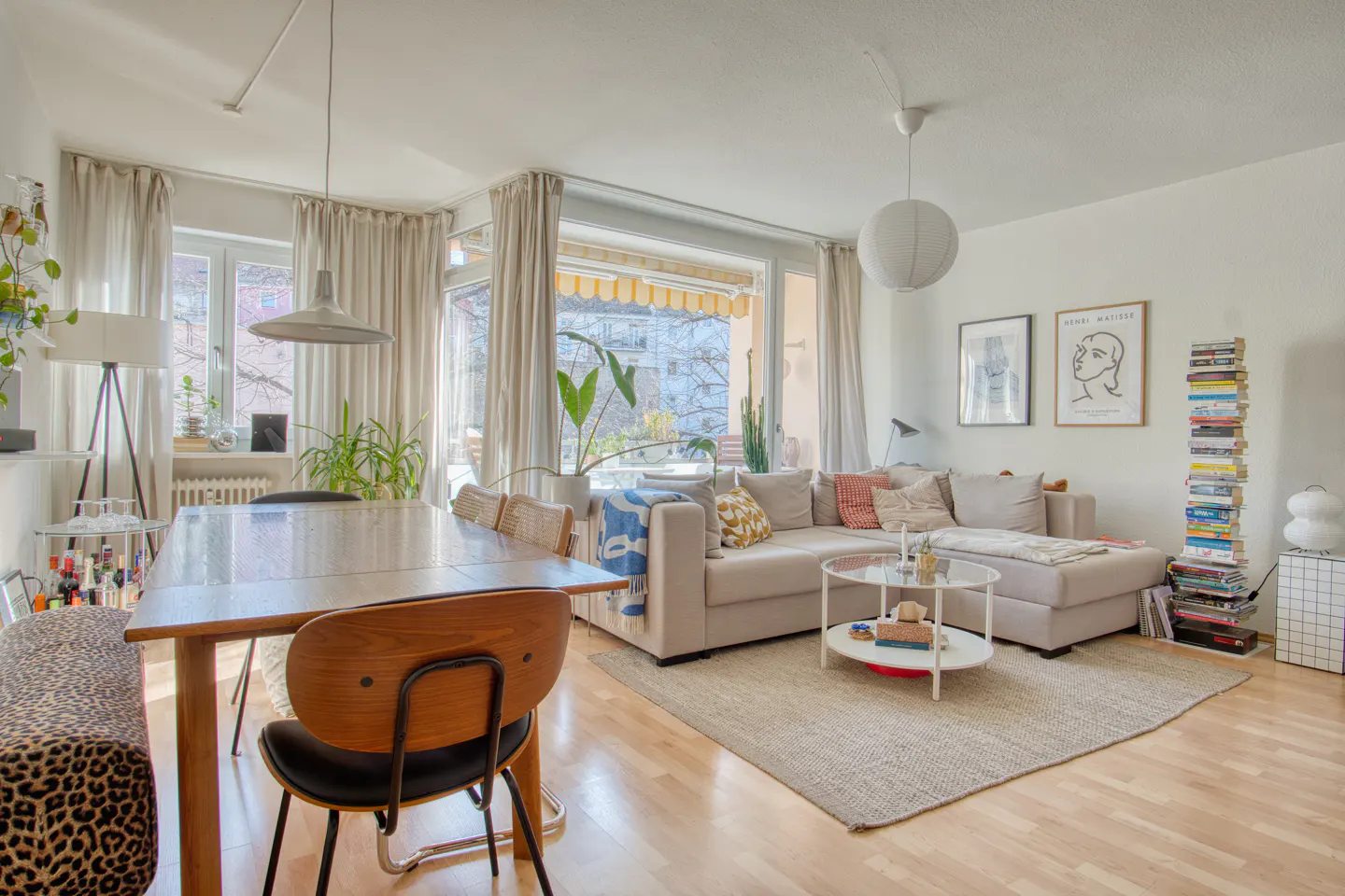Bright living room with a wooden dining table, beige sectional sofa, and light hardwood floors. Artwork and books add personality.