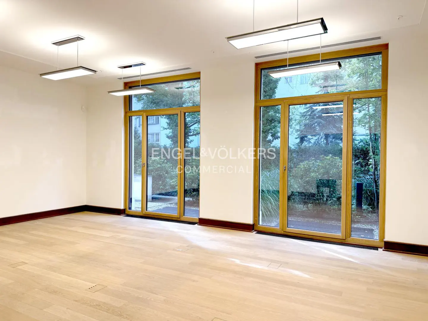 Bright, empty room with light wood floors, white walls, and large wood-framed windows overlooking greenery. Modern, rectangular light fixtures hang from the ceiling.