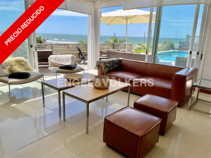 Bright living room with brown leather sofa, ottomans, and tables. Ocean view with pool and beach visible through large windows. "Precio Reducido" banner.