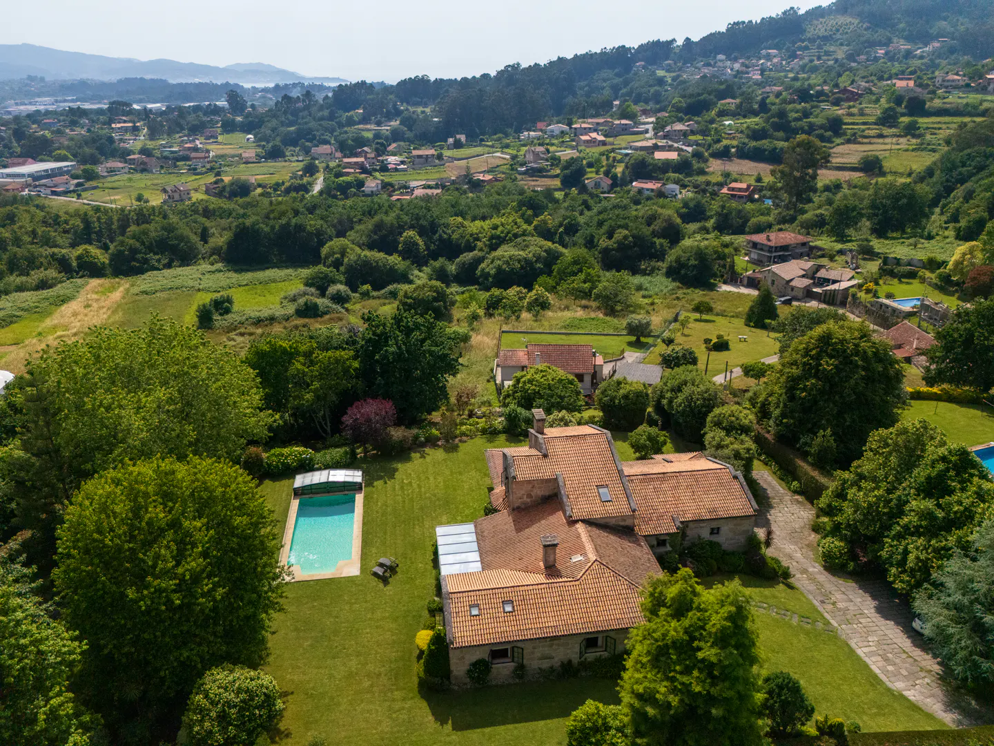 Aerial view of a stone house with a red tile roof, a pool, and green trees in a rural setting.