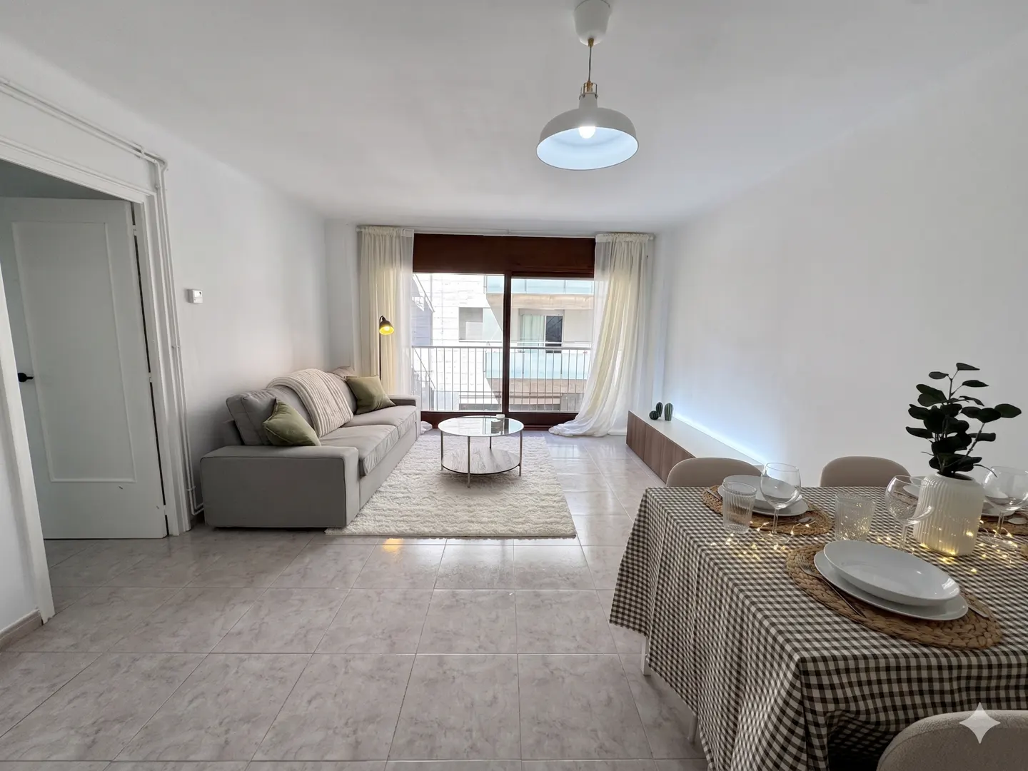 Bright living room with a gray sofa, round glass table on a white rug, and a dining table with a checkered tablecloth. Balcony in background.