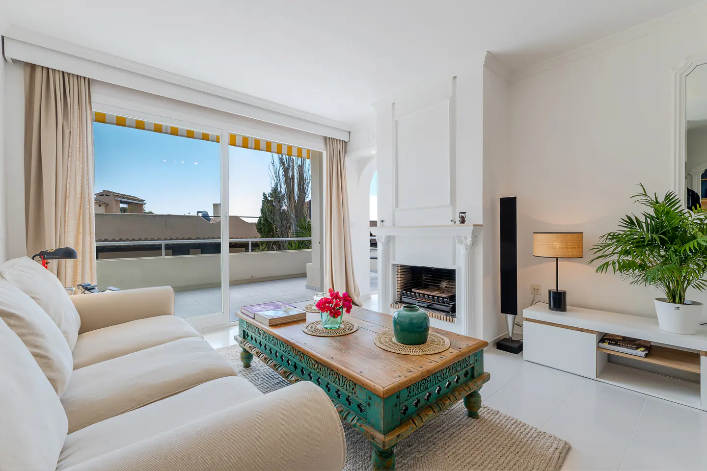 Bright living room with a beige sofa, carved wood coffee table, and white fireplace. Sliding glass doors lead to a balcony.