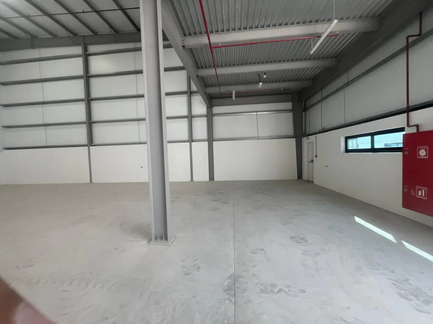Empty warehouse interior with gray concrete floor, white walls, and metal support beams. A red fire hose box is on the right wall.