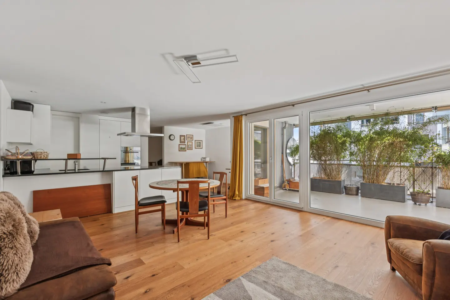 Bright, open-concept apartment with wood floors, a white kitchen, and a dining table. A plant-filled balcony is visible through sliding glass doors.