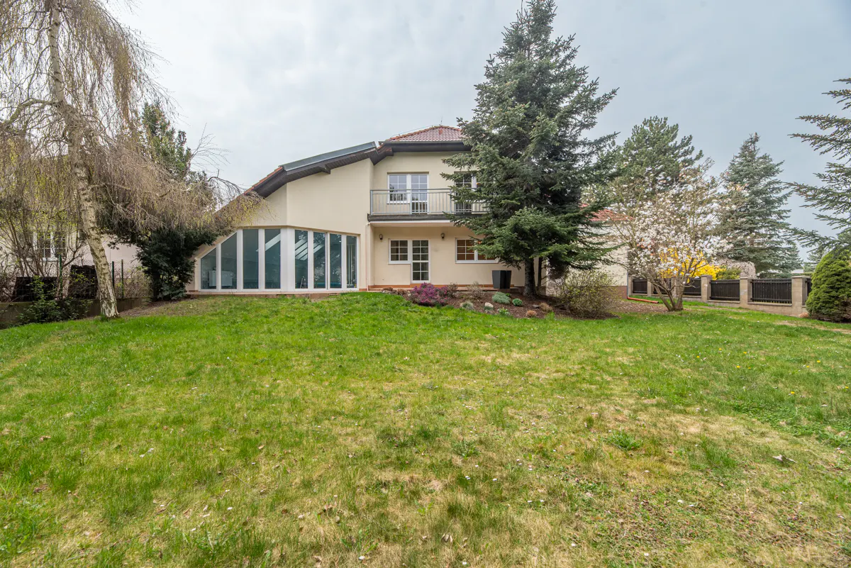 Two-story beige house with a red roof, a balcony, and a sunroom, surrounded by green grass and trees.