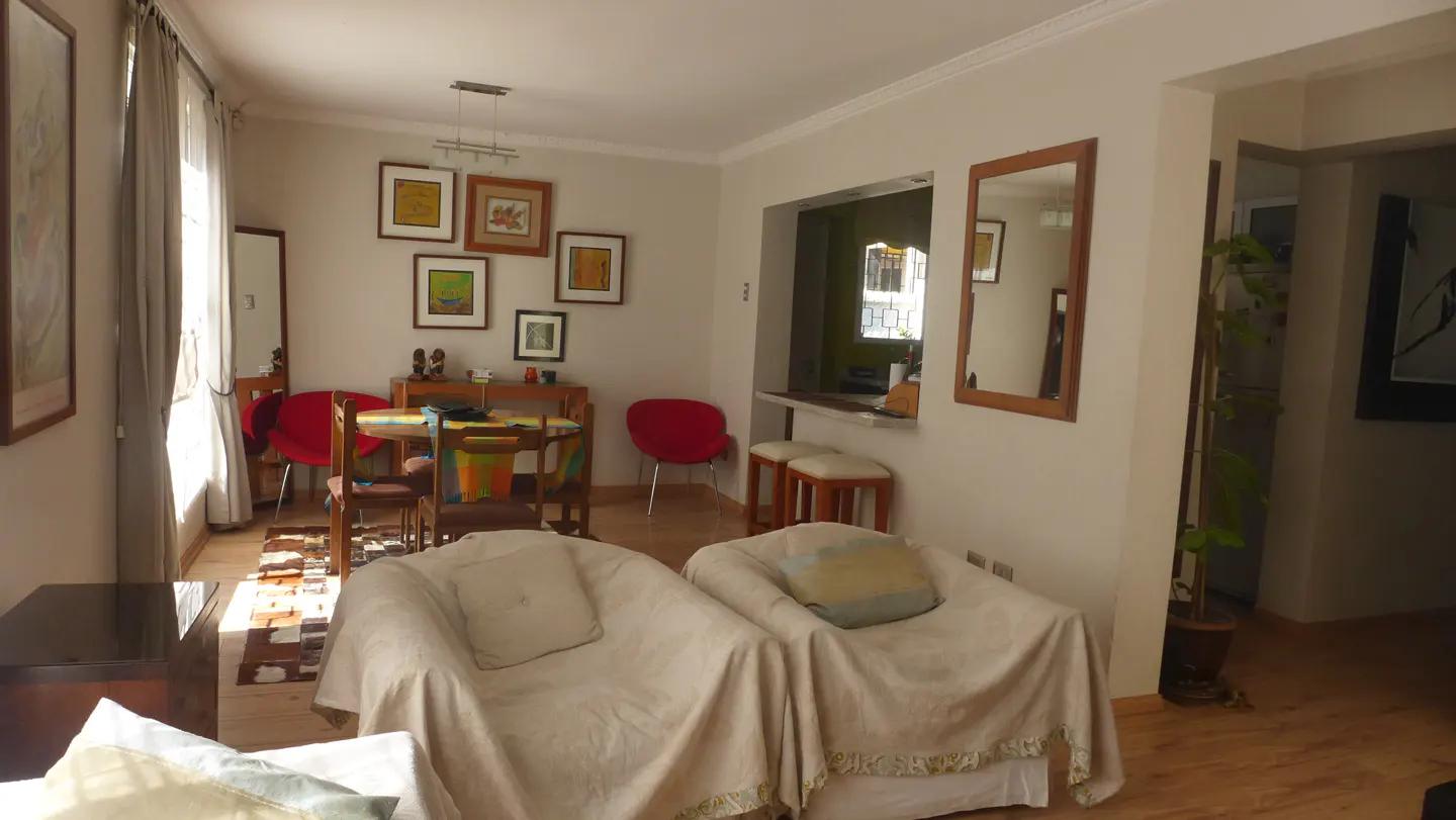 Living room with beige armchairs, wooden floors, dining table with red chairs, and framed art on the wall.