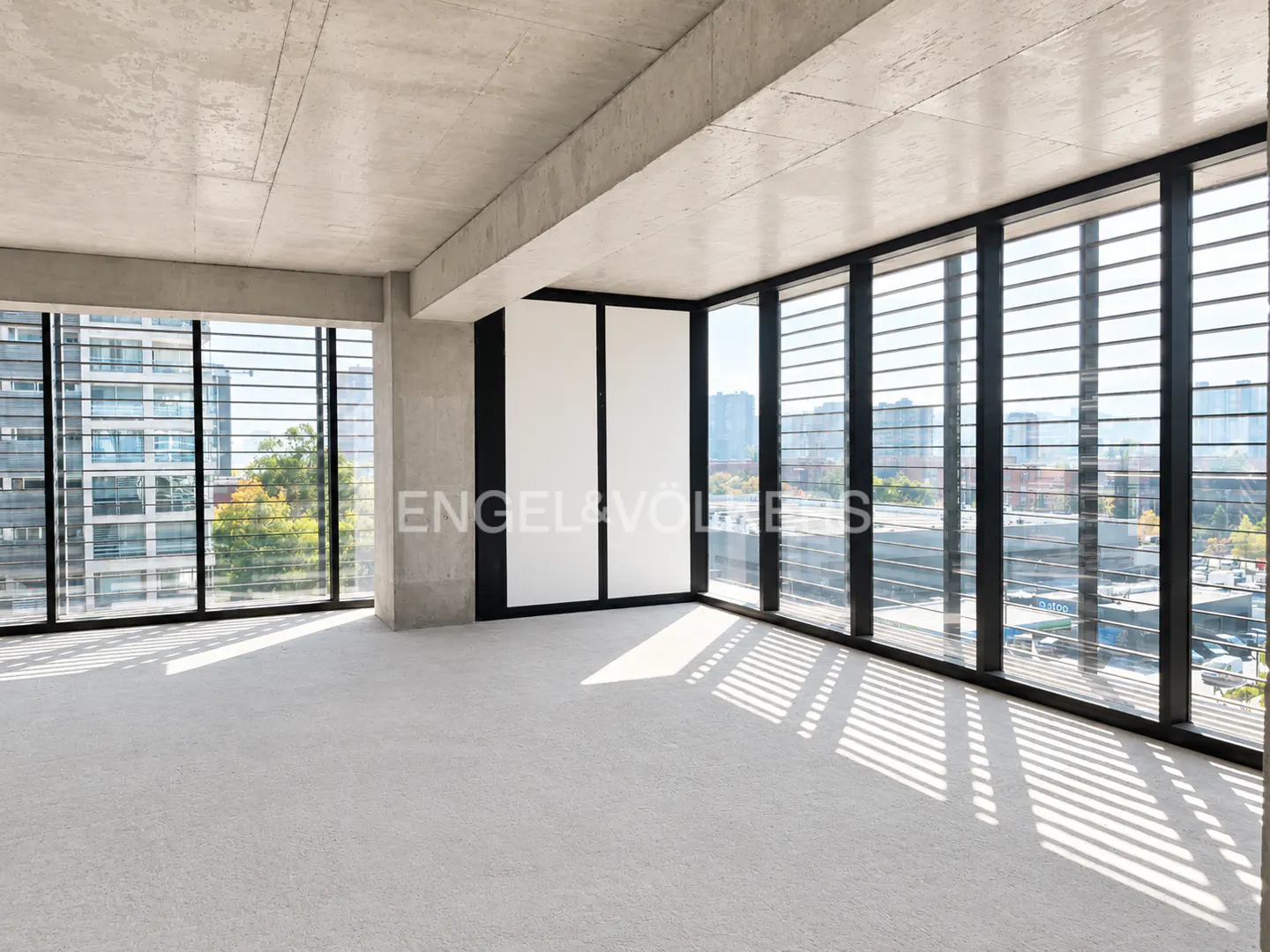 Bright, empty room with gray carpet, concrete ceiling, and black-framed windows with horizontal blinds overlooking a city.