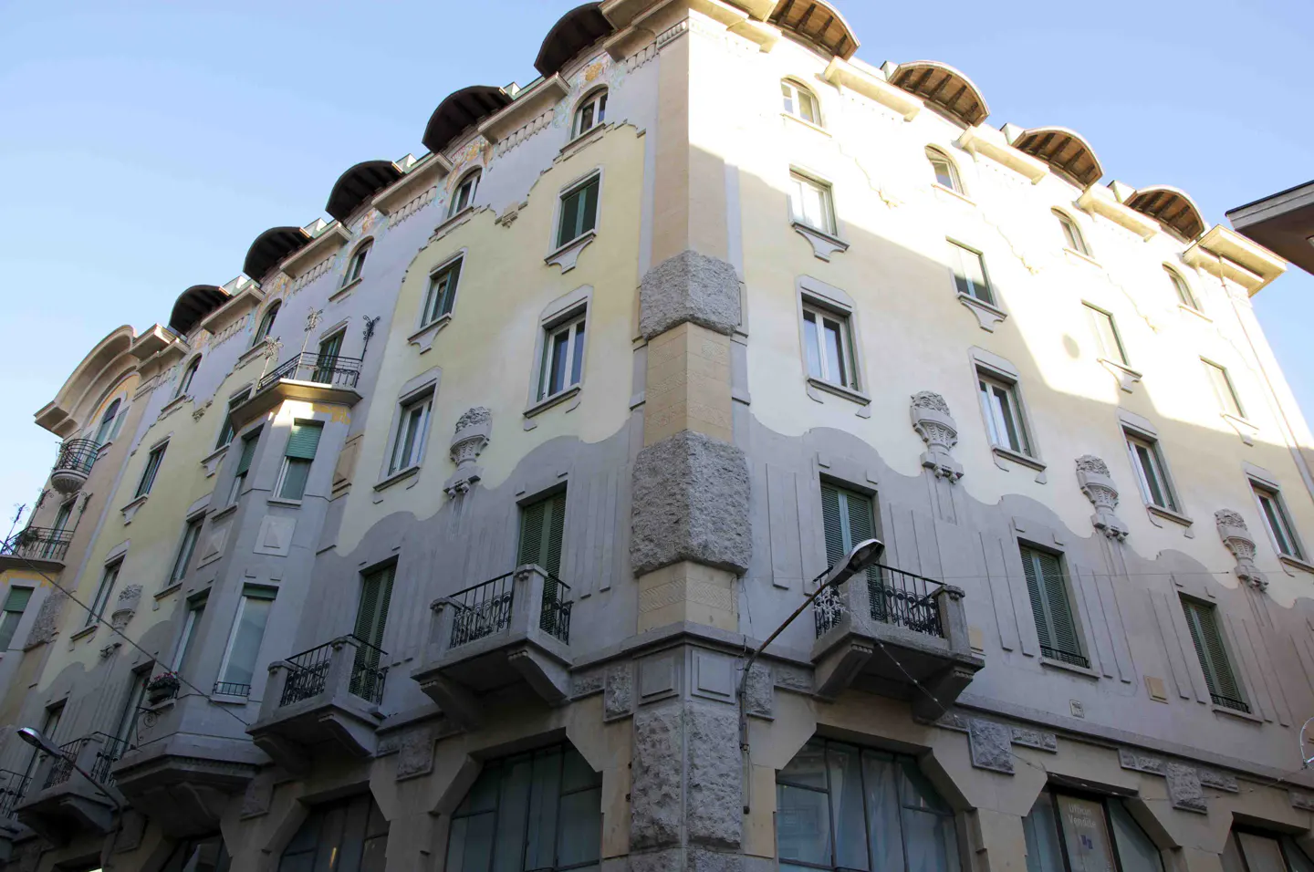 A low-angle shot of a multi-story building with a light yellow and gray facade, green windows, and small balconies.
